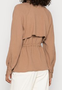 Bruine textuur top met een getailleerde taille, een gelaagde achterkant en lange mouwen die bij de manchet verzamelen. Licht tonale contrast tegen witte broeken.