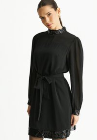 Black dress with long, sheer sleeves, a high collar, and a satin belt. Features a sequined hem and textured fabric.