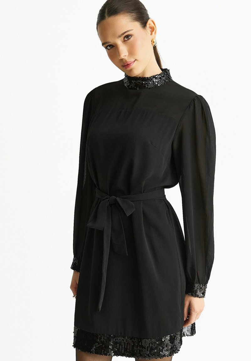 Black dress with long, sheer sleeves, a high collar, and a satin belt. Features a sequined hem and textured fabric.
