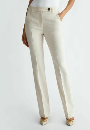 Woman wearing straight-cut beige trousers with front crease, side pockets, and a single black button, paired with beige pointed shoes with gold detail.