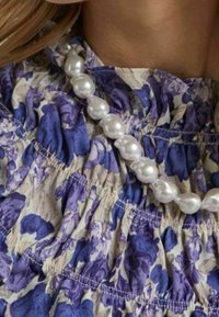 Blommönstrat tyg i nyanser av blått och gräddvit, med smockdetaljer och kompletterat med ett vitt pärlhalsband.