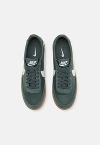 Sapatilhas de couro verde-escuro com o swoosh branco da Nike, atacadores texturizados e sola de borracha castanha. Apresentam interior acolchoado e logótipo na língua.