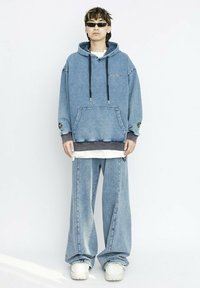 Sudadera de mezclilla con cordones, dos bolsillos frontales y logo; combinada con pantalones de mezclilla de corte holgado. El modelo lleva zapatillas blancas y gafas de sol.