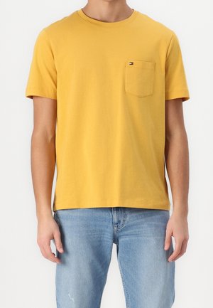 Homme portant un T-shirt jaune moutarde à manches courtes avec une poche poitrine et un jean bleu clair, debout devant un fond blanc uni.