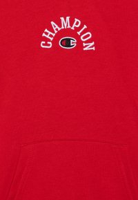 Sweatshirt en coton rouge avec une poche kangourou. Il présente un texte brodé blanc "CHAMPION" en arc au-dessus d'un logo noir et rouge. Texture lisse.