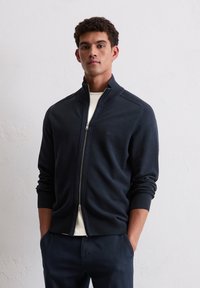 Marineblaue Zip-Up-Strickjacke aus strukturiertem Strickstoff, mit hohem Kragen und gerippten Bündchen. Minimales Logodesign auf der Brust.