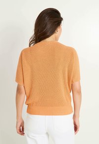 Top de punto naranja de manga corta con un corte relajado, textura acanalada y un dobladillo que se alinea con pantalones blancos de cintura alta. Sin cierres visibles.