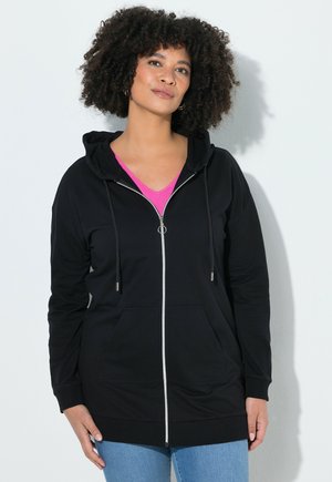 Vrouw met krullend haar die een zwart zip-up hoodie draagt over een roze shirt, staand tegen een lichtgrijze achtergrond, licht naar links kijkend.