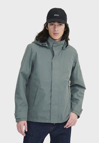 Veste à capuche résistant à l'eau en gris clair avec une texture lisse, fermeture à pression à l'avant et poches latérales. Portée avec une casquette noire.