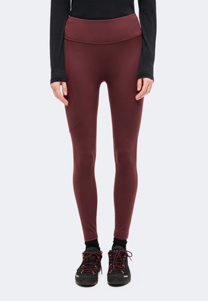 Modèle portant un legging bordeaux taille haute avec une poche latérale, un haut noir à manches longues et des chaussures de randonnée noires à lacets rouges.