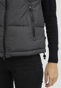 Gilet matelassé gris avec deux poches zippées diagonales, un col haut, un ourlet réglable et un tissu texturé, associé à un jean noir et un pull en maille.