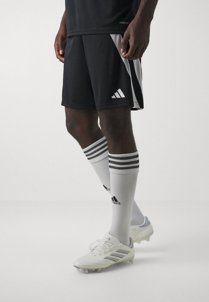 Shorts de sport noirs avec panneaux latéraux blancs, associés à des chaussettes blanches avec des rayures noires et le logo Adidas, ainsi que des crampons de football blancs.