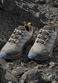 Paire de chaussures de randonnée grises et beiges avec lacets blancs reposant sur un terrain rocheux, dotée de semelles robustes et de languettes orange.