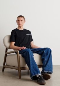 Black cotton t-shirt, front logo, relaxed fit. Light blue denim jeans, wide leg. Sitting on a beige upholstered chair with wooden legs.