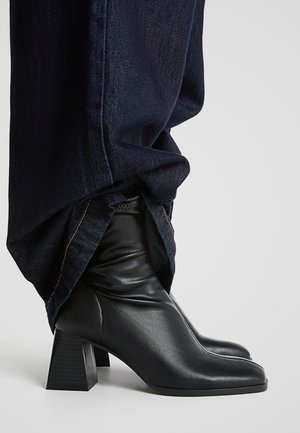 Schwarze Leder-Stiefeletten mit Blockabsätzen, getragen unter dunkelblauen Jeanshosen vor neutralem Hintergrund.