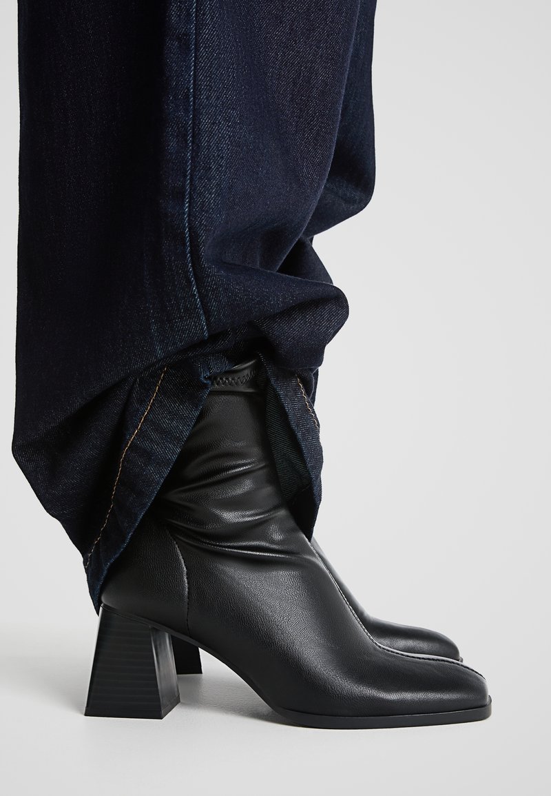 Bottines en cuir noir à talons blocs portées sous un pantalon en denim bleu foncé sur un fond neutre.