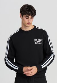 Felpa nera con accenti a strisce bianche sulle maniche e una grafica "New Jersey East Coast" sul petto. Tessuto morbido.