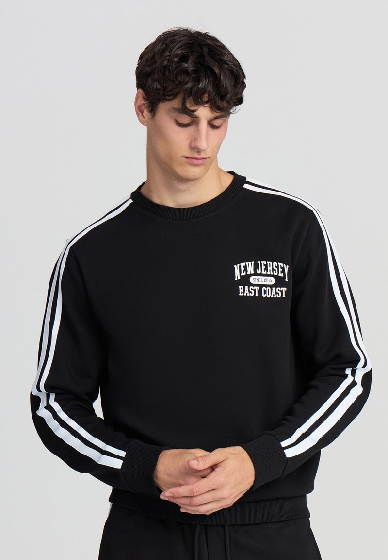 Felpa nera con accenti a strisce bianche sulle maniche e una grafica "New Jersey East Coast" sul petto. Tessuto morbido.