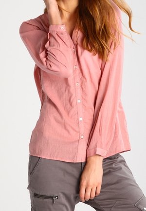 Femme portant une chemise rose à manches longues boutonnée et un pantalon cargo gris avec une poche zippée, posant avec une main près du visage.