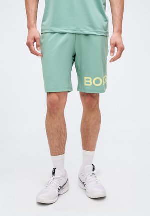 Muž nosí atletické šortky mintovej farby s textom „BORG“, biele ponožky a biele bežecké topánky, stojí na bielom pozadí.