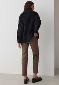 Veste noire surdimensionnée avec un col haut, texture mate et détails de boutons. Associée à un pantalon slim marron et des chaussures noires.