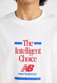 T-shirt de algodão branco com texto em vermelho e azul, dizendo "A Escolha Inteligente" e um logotipo "New Balance" em vermelho, com acessórios em azul.