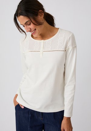 Donna sorridente e con lo sguardo rivolto verso il basso, indossa una camicia bianca a maniche lunghe con dettagli in pizzo e pantaloni blu scuro, mani nelle tasche, su sfondo semplice.