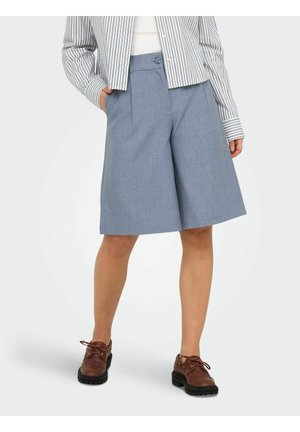 Kvinne som har på seg lyseblå, vide knelang shorts, stripete langermet skjorte og brune snøresko, står med hendene i lommene.