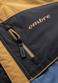 Gros plan d'une poche de veste noire et moutarde avec une fermeture éclair couleur moutarde et un curseur noir étiqueté "OMBRE."