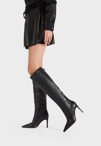 Stivali alti fino al ginocchio neri con tacco a stiletto realizzati in pelle liscia. Presentano una punta affusolata e un design aderente con un aspetto elegante e senza cuciture.