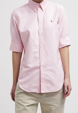 Chemise rose clair à boutons avec manches courtes, col pointu et petit logo brodé sur la poitrine gauche, portée avec un pantalon beige.