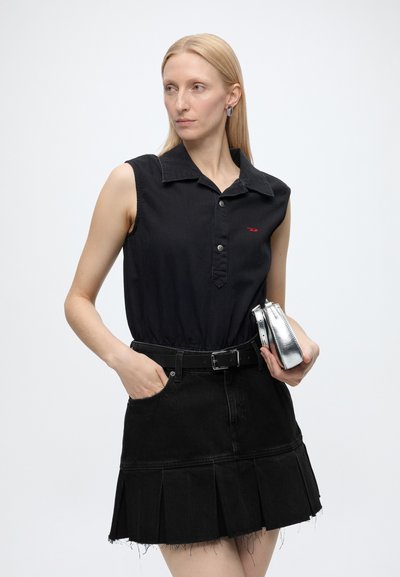 Femme aux longs cheveux blonds portant un haut noir sans manches à col et une jupe noire plissée, tenant une pochette argentée et regardant sur le côté.