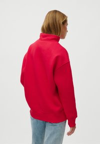 Pulover roșu cu guler înalt, croiala oversized, manșete și tiv ribbate. Materialul pare moale și are o textură netedă, fiind asortat cu blugi albastri deschiși.