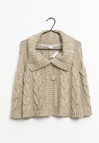 Cardigan beige tricoté avec un large col, orné de motifs torsadés, une fermeture par un bouton unique et des bords côtelés. Suspendu à un crochet noir.