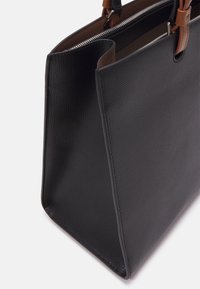 Czarna torebka tote z teksturowanej skóry z brązowymi uchwytami i widocznym srebrnym zamkiem wewnętrznym na białym tle.