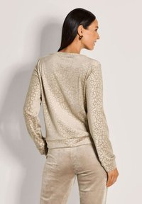 Vrouw met lang, donker haar, gekleed in een beige dierenprint met lange mouwen en bijpassende fluwelen broek, zijwaarts gekeerd met één hand in de zak.