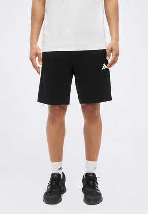 Mężczyzna w czarnych szortach Adidas, białych skarpetkach Adidas, czarnych butach sportowych Adidas i białej koszulce, stoi na jednolitym tle.