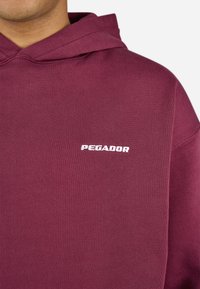 Maroon hoodie with hood and white "PEGADOR" logo embroidered on left chest, worn by a person against a plain background.