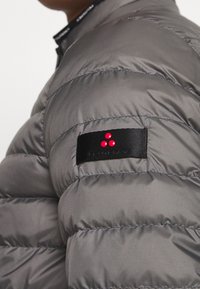 Grå pufferjakke med quiltet design, tekstureret stof og et sort mærke med tre røde prikker på skulderen.