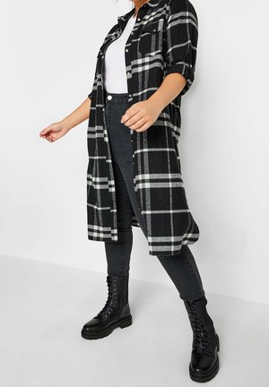 Persoon die een zwart-wit geruit lang overhemd draagt, een wit topje, zwarte skinny jeans en zwarte veter combat boots tegen een effen achtergrond.