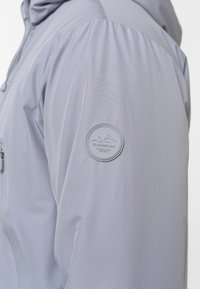 Lys grå udendørsjakke med tekstureret stof, med et rundt logo patch på ærmet og en lynlåslomme foran.