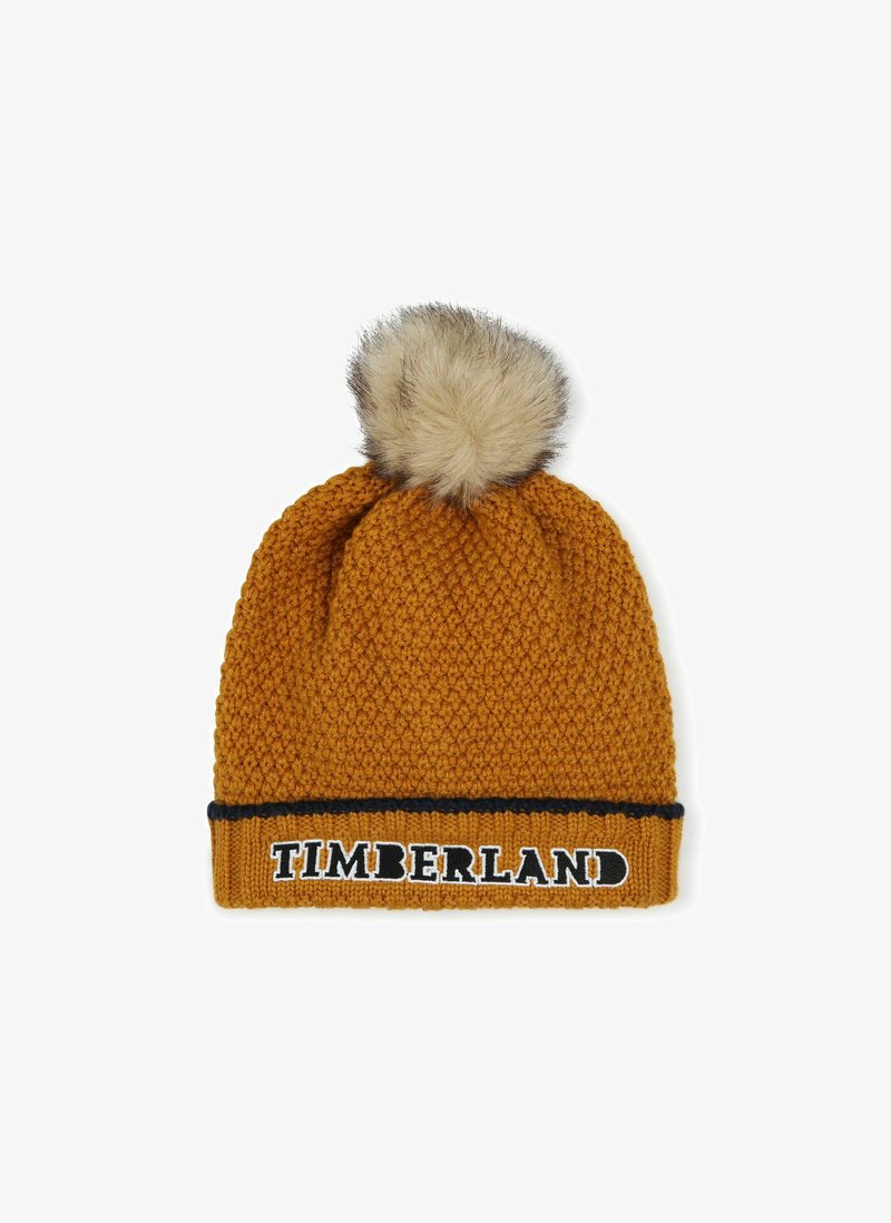 Bonnet tricoté en jaune moutarde avec un motif texturé, doté d'un pompon en fausse fourrure sur le dessus et d'une bande noire avec le logo "TIMBERLAND".