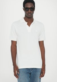 Hvit polo-skjorte med knapper i kragen, korte ermer og teksturert overflate. Har en liten sekskantet logo på brystet. Bært med blå jeans.