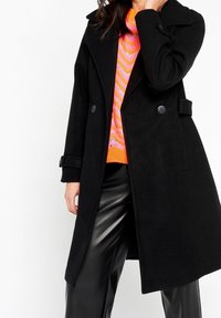 Czarny płaszcz z mieszanki wełny z dwurzędowym zapięciem, dużym kołnierzem i ozdobnymi guzikami. Połączony z jasnym, paskowanym swetrem i eleganckimi spodniami.