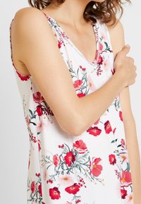 Donna che indossa un abito bianco senza maniche con stampa floreale rosa e rossa, incrociando un braccio sul petto contro uno sfondo neutro.