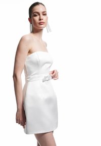 Weißes trägerloses Kleid mit einem taillierten Oberteil, einer Schleifendetail an der Taille und einer glatten Satin-Textur. Das Model trägt lange, mit Perlen besetzte Ohrringe.