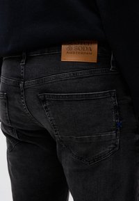 Κοντινό πλάνο ενός ατόμου που φοράει μαύρα τζιν με ένα καφέ δερμάτινο έμπλαστρο με την ένδειξη "Scotch & Soda Amsterdam" πάνω από την πίσω τσέπη.