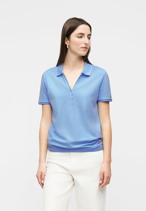 Kobieta ubrana w jasnoniebieską krótką koszulkę polo z białym obszyciem i białe spodnie, patrząca w bok na jednolitym tle.