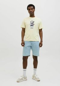 Lichtgele grafische t-shirt met een ontwerp van een schedel en vlinder, gecombineerd met lichtblauwe denim shorts. Witte sneakers met beige accenten en logo-sokken.