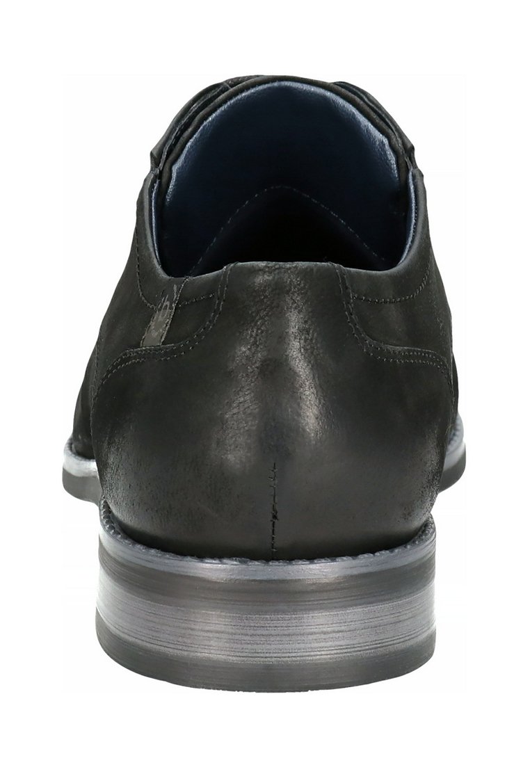 Zapato de vestir de cuero negro visto desde atrás, mostrando detalles de costura y una suela apilada de color gris.